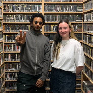 Toro y Moi and Maia Bilyk stand in the fbi music library facing the camera. Toro is wearing sunglassas and holding up his fingers in a peace sign to the camera. Maia is smiling next to him.