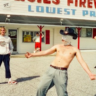 The five members of Shame stand in a gravel lot outside a large department stall. In the foreground one of the members is shirtless with a cowboy hat and foil over their face.