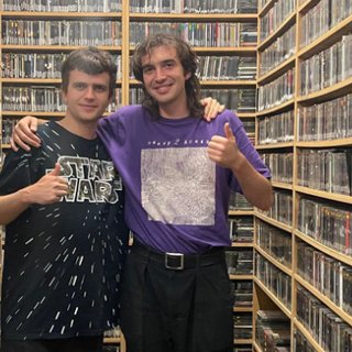 Eternal Dust’s guitarist Oscar Sulich and Jasper Craig-Adams stand in the fbi music library. They are facing the camera and smiling, with their arms around each others' shoulders. Both are doing a thumbs up.