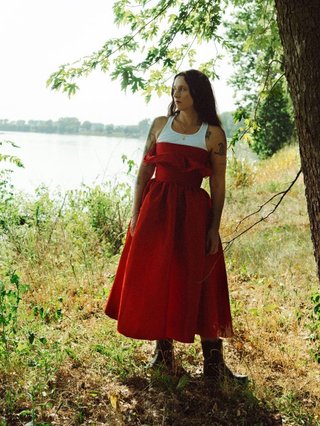 Waxahatchee. Woman in red dress standing by a river