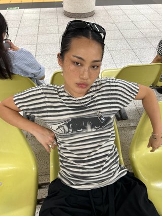 A high angle shot looking down at Yvonne. She's wearing a black and white photo looking into the camera with attitude. She appears to be in a public space, waiting on green chairs.