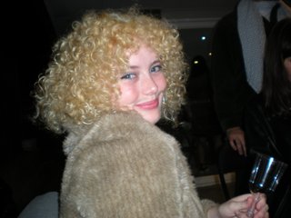 A digital camera photo of Zara looking to her right at the camera, smiling. She is wearing a light brown jumper and holding a wine glass.