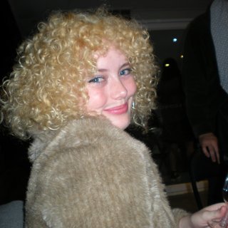 A digital camera photo of Zara looking to her right at the camera, smiling. She is wearing a light brown jumper and holding a wine glass.