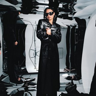 Erika de Casier poses at a photoshop wearing a long black trenchcoat and black sunglasses. The background is mirrored and stretched in psychedelic style