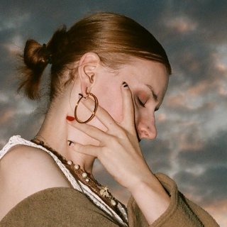 aya is standing against a cloudy sunset. She has her eyes closed and one hand covering her face.