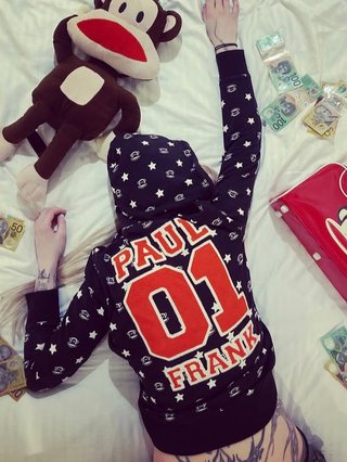 An image of Serena Blue laying face down on a bed surrounded by cash and stuffed toys.