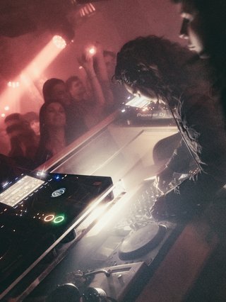 DJ at the club, slight crowd in view, light glowing from the DJ decks. Party is alive
