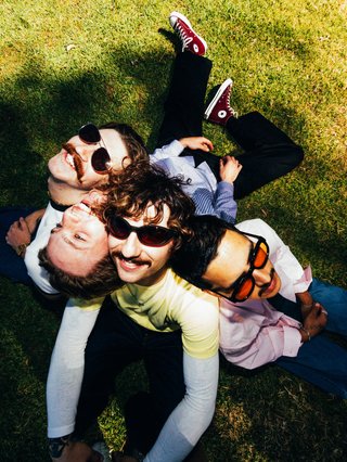 The four members of Perth indie-rock band DICE sit huddled together in the shade of a lush green lawn. The camera is angled down and they are all facing upwards to the camera, some with sunglasses. They are dressed in bright tones of chartreuse, pink, blue, burgundy, and deep denim. Their expressions are joyous and smiling.