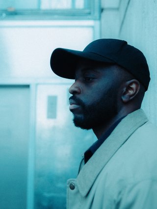 Alfa Mist in a black hat and light collared jacket facing side on. Photo has a cold, blue hue overtop to give it a filtered look.