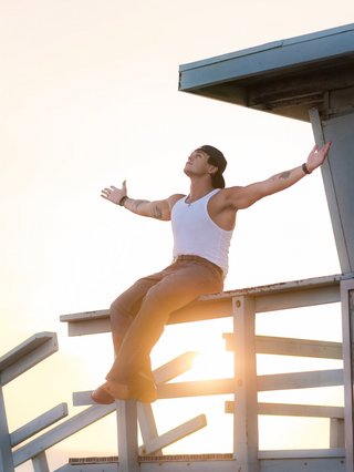Jonah Marais, a white man in jeans and a white tank top, sits posed on a railing with his arms outstretched. The sunset in the background saturates the image in pale yellow and blue tones.