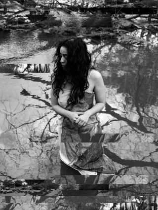 A black and white stylisted photo of sound artist Bint Mbareh standing knee deep in a pond with broken branches surrounding her. She is looking away as her hair sweeps over her face, the image is black and white equally thoughtful. The image was been glitched and collaged