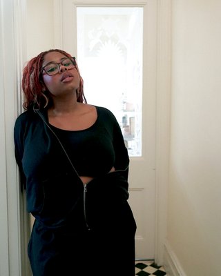 Ify stands in a white hallway leaning against a room door