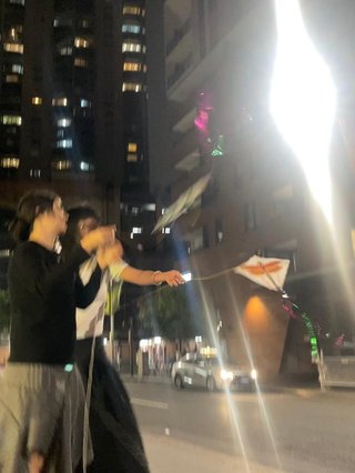 A blurry photo of Sehej and Wen Pei flying kites at night in the city. The photo is captured of them on their right side profile. The background is of city apartments and there is a light flash reflected on the top right corner of the photo.