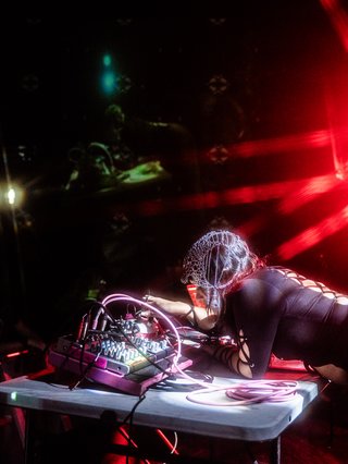 Carmen aka Society of Cutting Up Men (S.C.U.M) photographed in motion at a live set. She's crouched over her hardware and adorned in a chainmail headdress with a black dress.