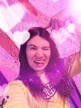 Jim making a cat ear pose, edited in front of a pink wall with lots of pink hearts and purple sparkles layered on top.