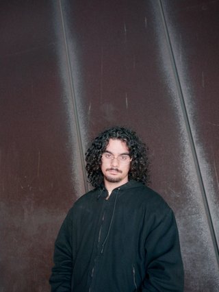 Miles Nautu stands in front of a red and grey striped wall. His curly dark hair is down to his shoulders, and he is wearing a navy windbreaker.