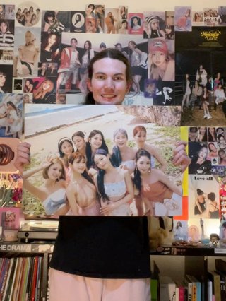 Jim standing in front of their wall of K-Pop posters, proudly holding up a poster of fromis_9