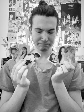 A black and white photo of Jim holding photocards of various K-Pop idols, including members of Cherry Bullet, cignature, fromis_9 and Loossemble. Jim has a sad expression on their face.