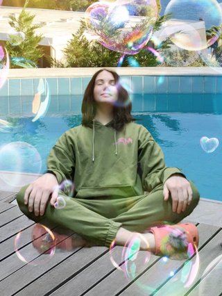 Jim sitting cross-legged wearing an olive green sweatsuit and orange Crocs. They have been edited in front of a pool scene, with bubbles floating around them.