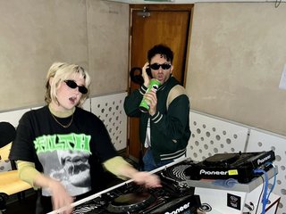 Ruby wears a Collarbones t shirt and pretends to play the DJ decks, Al sings into a pringles can. Both are wearing dark sunglasses in the FBi studio.