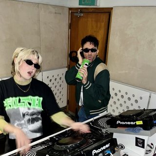 Ruby wears a Collarbones t shirt and pretends to play the DJ decks, Al sings into a pringles can. Both are wearing dark sunglasses in the FBi studio.