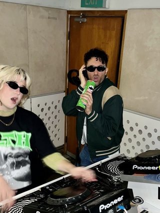Ruby wears a Collarbones t shirt and pretends to play the DJ decks, Al sings into a pringles can. Both are wearing dark sunglasses in the FBi studio.