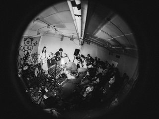 A fish eye black and white photo of a Garage Sale concert. The band is playing on stage in a small cramped room with a big crowd.