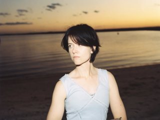 Georgia Mulligan is shot waist up, wearing a grey-blue singlet, looking to the left of frame. She is in front of a body of water at sunset