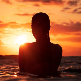 A girl standing in the ocean as waist level, staring into the rise of the sun.