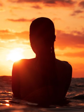 A girl standing in the ocean as waist level, staring into the rise of the sun.