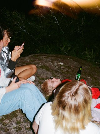 flash photo of 5 people sitting on a rock all facing behind their backs to the camera. theres an orange light flare on the right side of the photo and everyone is holding drinks