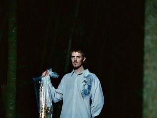 heartholder outside stands against a black background. Looking off to the side, his left arm is behind his back and his right arm holds his saxophone in a clear plastic bag