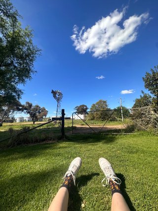 A POV view of the countryside on a sunny day, a wire fence, windmill and pair of legs wearing flame socks and white sneakers on the grass are visible