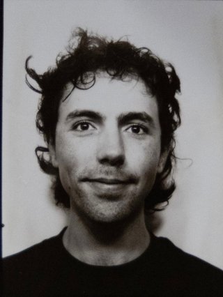 Black and white film photo booth photo of Mike who is looking directly into camera smiling. he is wearing a black shirt and has short curly black hair