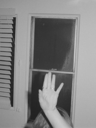 black and white photo of someone holding their hand to the air. you can see two windows in the background, one with white blinds over it. the image is cropped so you cant see the person's face, only the top of their head.