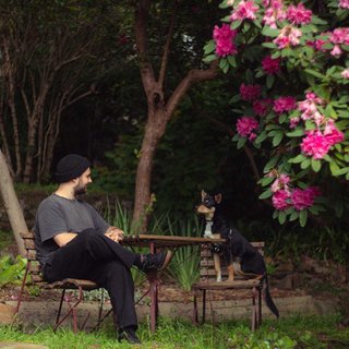 Jackson sits at an outdoor table, legs crossed and gazing at a black and brown dog also sitting at the table. They are in a lush, leafy setting with pink flowers.