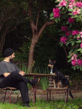Jackson sits at an outdoor table, legs crossed and gazing at a black and brown dog also sitting at the table. They are in a lush, leafy setting with pink flowers.