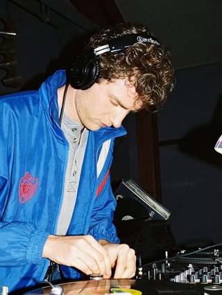 Luke is DJing wearing a set of black headphones and a shiny blue sports jacket