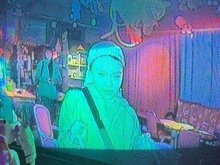 Sim is bathed in a green glow and seen through a television screen. They are looking down wearing a bandana and a bag strapped across their shoulder.