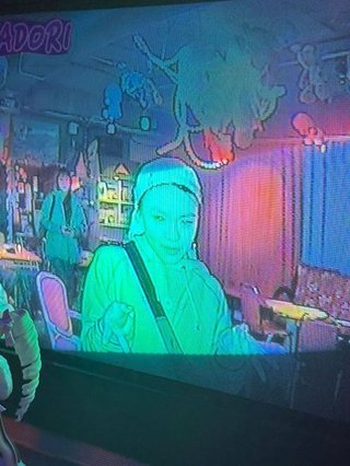 Sim is bathed in a green glow and seen through a television screen. They are looking down wearing a bandana and a bag strapped across their shoulder.
