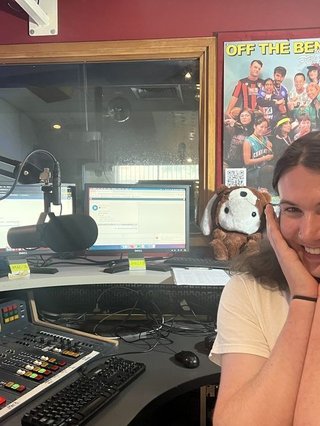 Jim is looking gleeful in the FBi on air studio, they are sitting in front of the mic, computers and panel with their hands cradling their chin and a smile on their face.