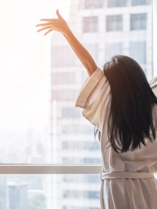 A stock image of a person with long black hair stretching in front of a sunny window. they are wearing a white robe