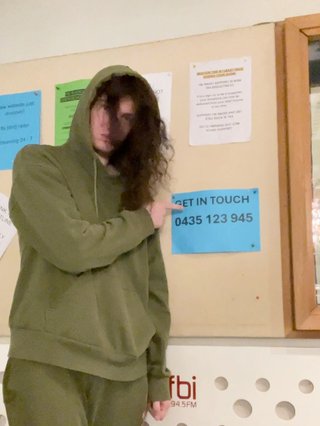Jim in an army green tracksuit with the hood on pointing at a sign in the studio that says "Get In Touch 0435123945"