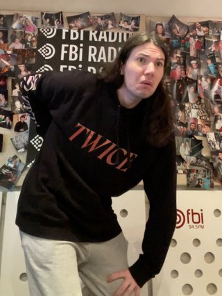 Jim standing in the fbi studio, hunched over with one hand on their knee and the other on their back as they pull a silly face.