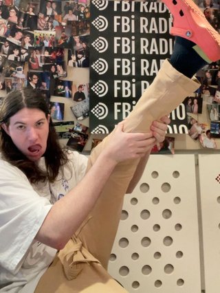 A photo of Jim in the fbi studio sitting on a chair and awkwardly holding their leg in the air, showing off their bright orange Crocs.