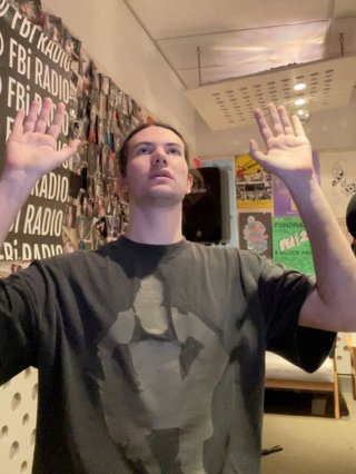 Jim in the fbi studio, holding their hands up beside their head.