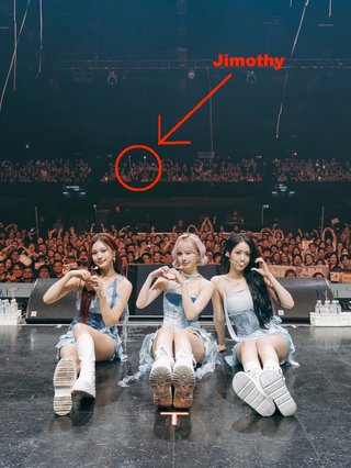 A photo of girl group VIVIZ from their recent Melbourne show. The picture features them sitting on the stage with the crowd in the background. In the very back of the crowd a small blur is highlighted by a red circle with an arrow labelled "Jimothy" pointing at it.
