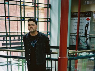 A full body portrait shot of now always fades standing inside a glass windowed, mall like building. He is wearing all black, and leaning against a railing.