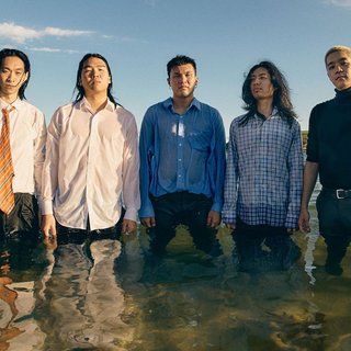 The five members of 1300 stand in thigh-deep water facing the camera. They are at the beach and are all wet and wearing shirts and suit pants.
