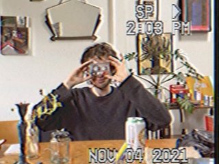 Jasper Craig Adams holds a vintage camera to his eye, obscuring his face. He is wearing a grey jumper, sitting at a wooded dining table where an assortment of objects are placed - a candleholder, a jar of plant cuttings, a can of drink, and some papers. Behind him a white wall features various artworks and a hanging mirror. The image is captured as if from a digital camera still, complete with slight pixelation and stamped with the time and date with white digital text